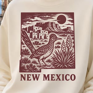 May include: A cream-colored crewneck sweatshirt featuring a maroon graphic print of a roadrunner, desert landscape, and the text NEW MEXICO. The design includes a sun, mesas, and cacti, centered on the chest of the long-sleeved pullover garment.