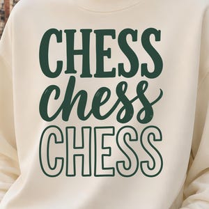 May include: A cream-colored crewneck sweatshirt featuring the word CHESS printed three times in dark green. The first line is in a bold serif font, the second in a cursive script, and the third in a hollow outline font. The sweatshirt has long sleeves.