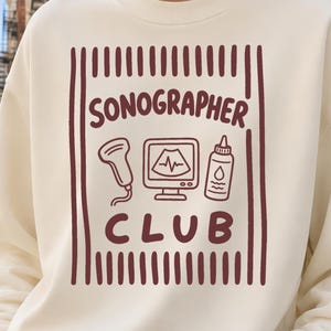 May include: A cream-colored crewneck sweatshirt featuring a dark red graphic print. The design includes vertical stripes, the text "SONOGRAPHER CLUB," and icons of an ultrasound probe, a monitor displaying a waveform, and a bottle of ultrasound gel.