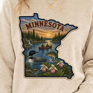 May include: A beige crewneck sweatshirt featuring a large, detailed graphic shaped like the state of Minnesota. The design depicts a sunset over a lake with a canoe, a loon, a jumping fish, pine trees, and pink lady slipper flowers. The text MINNESOTA is printed in a bold, dark red font at the top of the state-shaped graphic.
