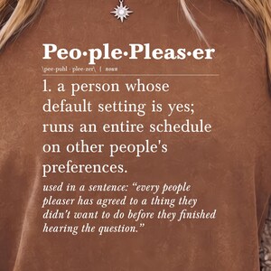 Op de afbeelding: Een bruin t-shirt met korte mouwen en witte tekst die het woord people-pleaser definieert als een persoon wiens standaardinstelling ja is, die een volledig schema beheert op basis van de voorkeuren van anderen, inclusief een voorbeeldzin.