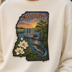 May include: A cream-colored crewneck sweatshirt featuring a graphic shaped like the state of Mississippi. The design includes a paddlewheel boat on a river, a jumping fish, magnolia flowers, and the text MISSISSIPPI under a sunset sky.