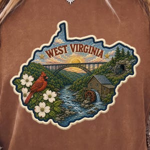May include: A brown t-shirt featuring a graphic shaped like the state of West Virginia. The design includes a cardinal, white dogwood flowers, a river, a water mill, a bridge, and a sunset. The text "WEST VIRGINIA" is printed at the top.