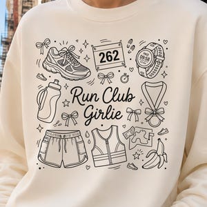 Op de afbeelding: Cr&egrave;mekleurige sweatshirt met een zwart-wit grafisch ontwerp met hardloop-thema illustraties en de tekst "Run Club Girlie". Het ontwerp bevat een hardloopschoen, shorts, een waterfles en een horloge.