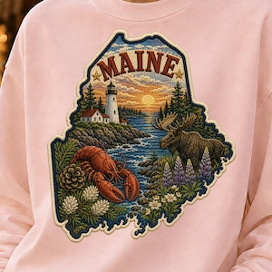 Pode incluir: Uma camisola rosa claro com um gráfico grande e detalhado do estado do Maine. O design inclui um farol, um alce, uma lagosta, pinheiros e flores de tremoço, com a palavra MAINE impressa em letras vermelhas arrojadas no topo.