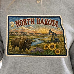 May include: A heather grey crewneck sweatshirt featuring a rectangular graphic of the state of North Dakota. The design includes a brown bison, a winding river, a black oil pump jack, sunflowers, and a sunrise, with the text NORTH DAKOTA.