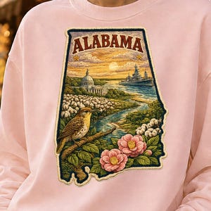 May include: A light pink crewneck sweatshirt featuring a large, detailed graphic shaped like the state of Alabama. The design includes the text ALABAMA, a bird on a branch, pink camellia flowers, cotton plants, a domed building, and a ship.