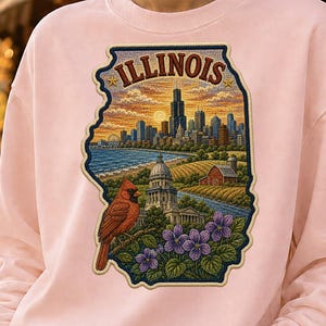 Peut inclure: Un sweat-shirt à col rond rose clair arborant un grand motif en forme de l'État de l'Illinois. Le design comprend un paysage urbain détaillé, un cardinal rouge perché sur une branche, des violettes, une ferme et le texte ILLINOIS en haut.