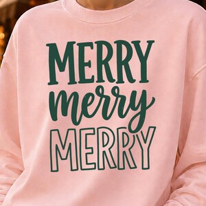Könnte beinhalten: Ein hellrosa Sweatshirt mit Rundhalsausschnitt und dem in Dunkelgrün auf die Brust gedruckten Text MERRY merry MERRY. Das Sweatshirt hat lange Ärmel und einen gerippten Ausschnitt und wird mit einer blauen Jeans im klassischen Five-Pocket-Stil kombiniert.