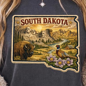 May include: A charcoal grey t-shirt featuring a rectangular graphic of South Dakota. The design includes Mount Rushmore, a bison, a pheasant, and purple pasqueflower blossoms set against a landscape with a river, mountains, and a small barn.