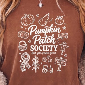 Peut inclure: T-shirt marron avec un motif blanc « Pumpkin Patch Society » orné de citrouilles, d'un épouvantail et d'autres éléments d'automne. Le t-shirt à col rond est de couleur unie.