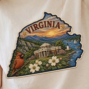 May include: A beige t-shirt featuring a large, detailed graphic shaped like the state of Virginia. The design includes a red cardinal, white dogwood flowers, a historic mansion, a lighthouse, sailboats, and mountains under a sunset sky.