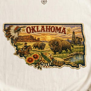 May include: A cream-colored t-shirt featuring a large, detailed graphic shaped like the state of Oklahoma. The design includes a cowboy on horseback, bison, sunflowers, a drum, an oil derrick, and a Native American headdress with the text OKLAHOMA.