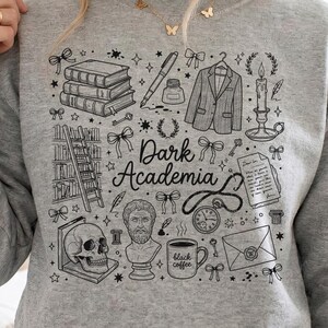 May include: A grey sweatshirt with a black graphic design. The design includes books, a pen, a jacket, a candle, a skull, a bust, and the words "Dark Academia". The design also features stars and bows.
