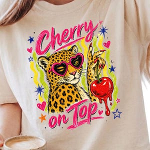 Op de afbeelding: Beige T-shirt met een luipaard met een hartvormige zonnebril en een kers. De tekst "Cherry on Top" is in het roze geschreven. Het ontwerp bevat sterren, harten en een gele en groene golvende achtergrond.