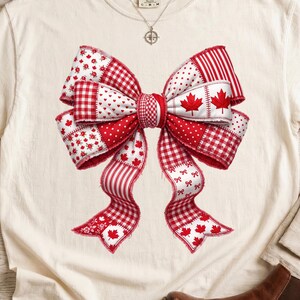 May include: Cream-coloured t-shirt with a large bow design. The bow is a patchwork of red and white patterns, including gingham, hearts, and maple leaves. The shirt has a round neckline and short sleeves.