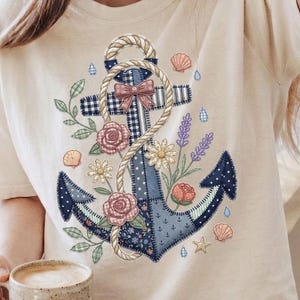 Può includere: T-shirt color crema con un design di ancora nautica. L'ancora è decorata con motivi floreali e patchwork, tra cui rose, margherite e un fiocco a quadretti. Una corda avvolge l'ancora e conchiglie e gocce d'acqua sono sparse intorno al design.