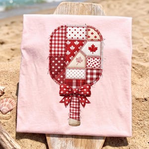 May include: A light pink t-shirt featuring a decorative design of a popsicle with a patchwork pattern in red and white. The popsicle design includes maple leaves and a bow at the base. The shirt is displayed on a wooden surface near the beach.