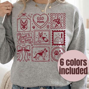 May include: Gray sweatshirt with a maroon graphic featuring German Shepherd illustrations, paw prints, and the text "GSD Mom" and "Loyal To The Bone." A pink circle with the text "6 colors included" is also visible.
