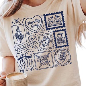 May include: A cream-coloured t-shirt with a dark blue design. The design includes a heart with the text "Morally Grey", a bow, a sword through a rose, and the text "Who Did This To You".