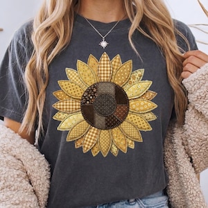 May include: Dark gray t-shirt featuring a large sunflower design. The sunflower has a patchwork design with various yellow and brown patterns. The shirt is a casual style, suitable for everyday wear.