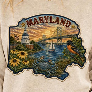 May include: A beige crewneck sweatshirt featuring a graphic print of the state of Maryland. The design includes a blue crab, a Baltimore oriole, sunflowers, a sailboat, a bridge, and a building, with the text MARYLAND at the top.