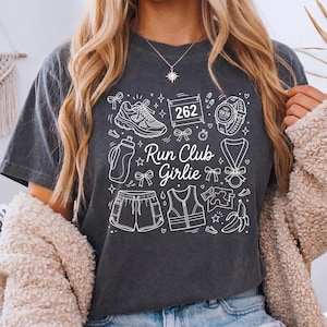 Op de afbeelding: Donkergrijs t-shirt met een wit grafisch ontwerp met hardloopgerelateerde iconen en de tekst "Run Club Girlie". Het ontwerp bevat een hardloopschoen, waterfles, korte broek, vest, horloge en een medaille.