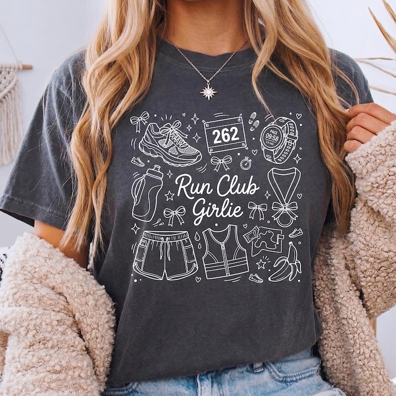 Op de afbeelding: Donkergrijs t-shirt met een wit grafisch ontwerp met hardloopgerelateerde iconen en de tekst "Run Club Girlie". Het ontwerp bevat een hardloopschoen, waterfles, korte broek, vest, horloge en een medaille.