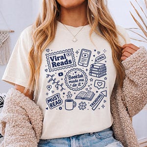 May include: Cream-colored t-shirt with a navy blue graphic design featuring the words "Viral Reads," "Booktok made me do it," and "Must Read." The design includes book-related icons, a phone, and the word "Trending."