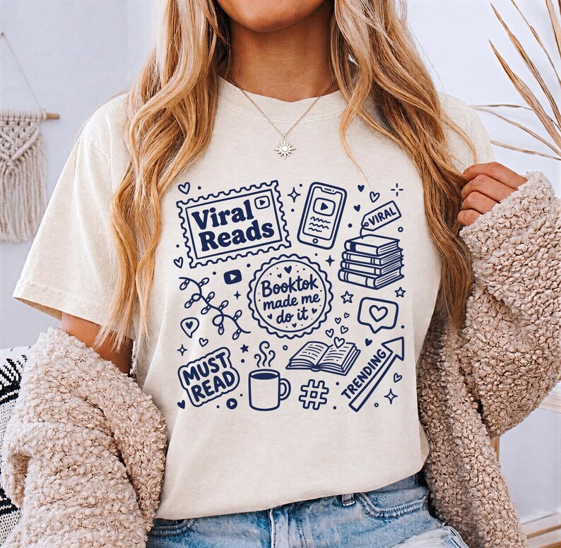 May include: Cream-colored t-shirt with a navy blue graphic design featuring the words "Viral Reads," "Booktok made me do it," and "Must Read." The design includes book-related icons, a phone, and the word "Trending."