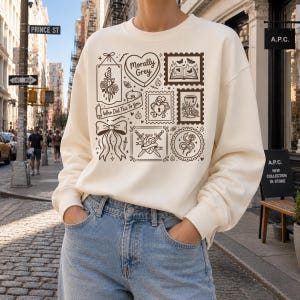 May include: Cream-colored sweatshirt with a collection of brown graphic illustrations. The design includes a heart, a bow, a sword, a snake, and the text "Morally Grey" and "Who Did This To You". The sweatshirt is paired with blue jeans.