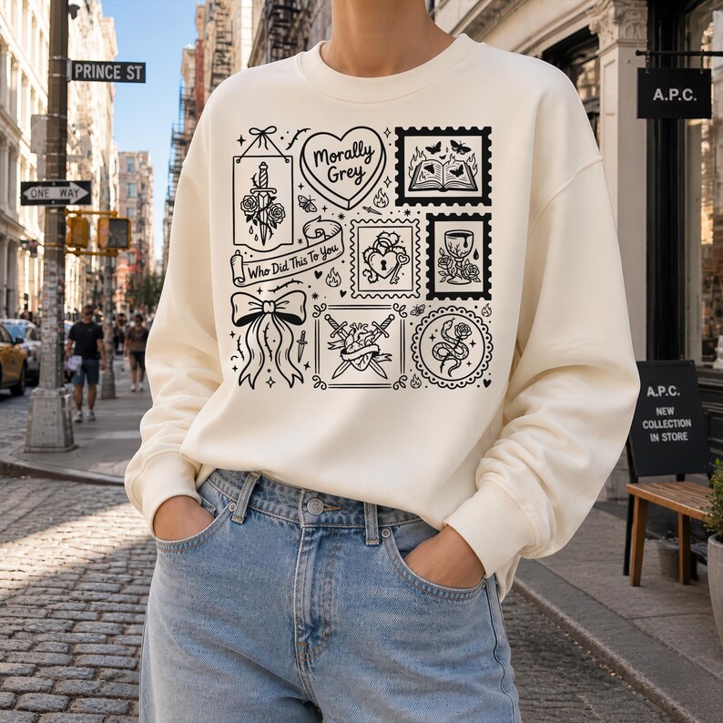 May include: Cream-colored sweatshirt with a black graphic design featuring various illustrations, including a heart, bow, and sword. The text "Morally Grey" and "Who Did This To You" are also included in the design. The person is wearing blue jeans.