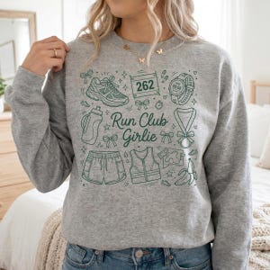 Op de afbeelding: Een grijze sweatshirt met een groene grafische opdruk met hardloopgerelateerde illustraties. Het ontwerp bevat een hardloopschoen, een waterfles, een korte broek, een vest, een marathonnummer, een horloge, een medaille en een banaan. De tekst "Run Club Girlie" is ook inbegrepen.