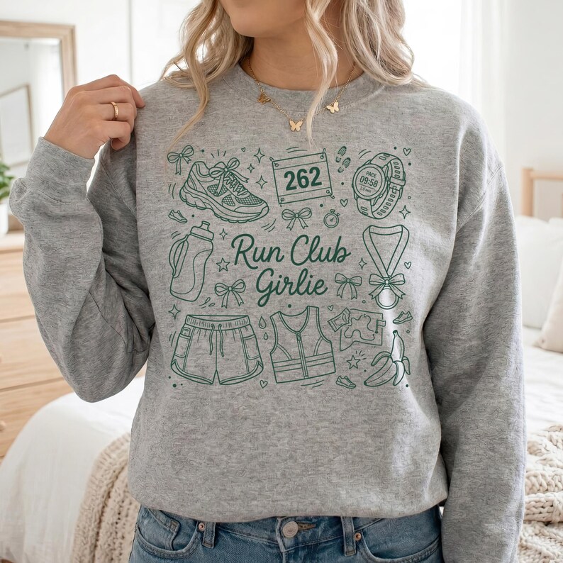Op de afbeelding: Een grijze sweatshirt met een groene grafische opdruk met hardloopgerelateerde illustraties. Het ontwerp bevat een hardloopschoen, een waterfles, een korte broek, een vest, een marathonnummer, een horloge, een medaille en een banaan. De tekst "Run Club Girlie" is ook inbegrepen.