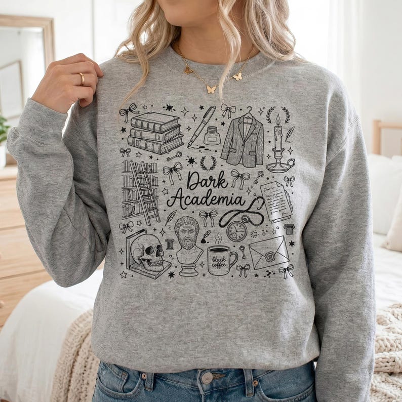 May include: A grey sweatshirt with a black graphic design. The design includes books, a pen, a jacket, a candle, a skull, a bust, and the words "Dark Academia". The design also features stars and bows.