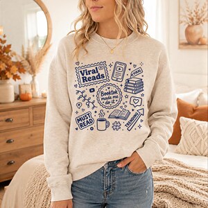 May include: A light beige sweatshirt with a blue graphic design featuring the words "Viral Reads," "Booktok made me do it," and other book-related icons. The design includes hearts, a coffee cup, and a trending arrow.