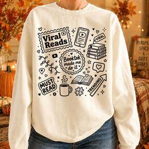 May include: Cream-colored sweatshirt with black graphic illustrations related to reading and social media. The design includes the text "Viral Reads," "Booktok made me do it," and "Must Read," along with icons of books, a phone, and a coffee cup.