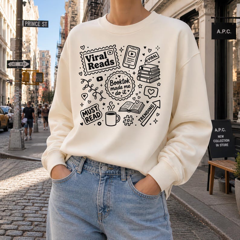 May include: Cream-colored sweatshirt with a black graphic design featuring the words "Viral Reads," "Booktok made me do it," and "Must Read." The design includes book stacks, a coffee mug, a phone, and other book-related icons. The person is wearing blue jeans.