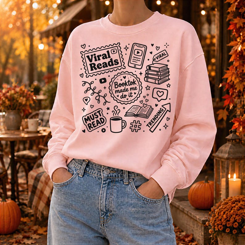 May include: A light pink sweatshirt with a black graphic design featuring the words "Viral Reads", "Booktok made me do it", and "Must Read". The design also includes book and social media icons. The person is wearing blue jeans.
