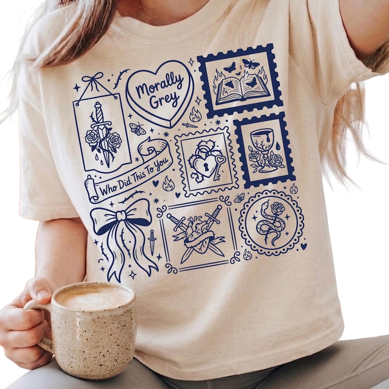 May include: A cream-coloured t-shirt with a dark blue design. The design includes a heart with the text "Morally Grey", a bow, a sword through a rose, and the text "Who Did This To You".