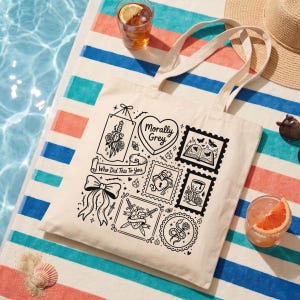 May include: Cream-colored tote bag with black graphic illustrations and text. The bag features a variety of designs, including a heart with the words "Morally Grey", a sword through a rose, and a bow. The text "Who Did This To You" is also present.