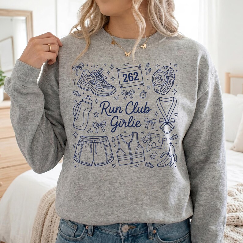 Op de afbeelding: Een grijze sweatshirt met een blauw grafisch ontwerp. Het ontwerp bevat hardloopschoenen, een waterfles, een korte broek, een vest, een startnummer, een horloge, een medaille en een banaan. De tekst "Run Club Girlie" is ook inbegrepen.