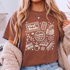 May include: A brown t-shirt with white graphics related to reading and social media. The shirt features the text "Viral Reads", "Booktok made me do it", "Must Read", and "Trending", along with icons of books, phones, and coffee.