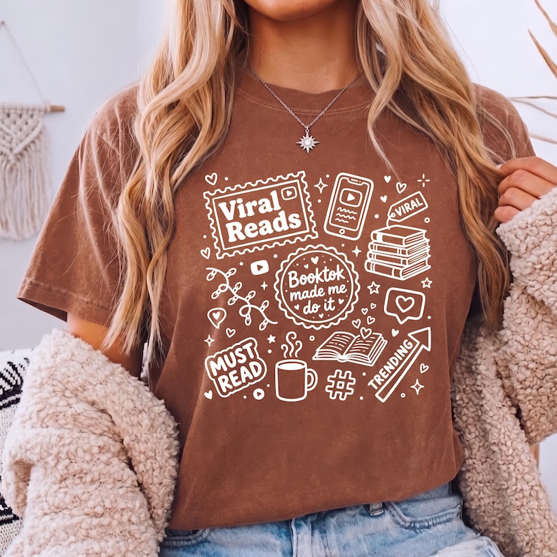 May include: A brown t-shirt with white graphics related to reading and social media. The shirt features the text "Viral Reads", "Booktok made me do it", "Must Read", and "Trending", along with icons of books, phones, and coffee.
