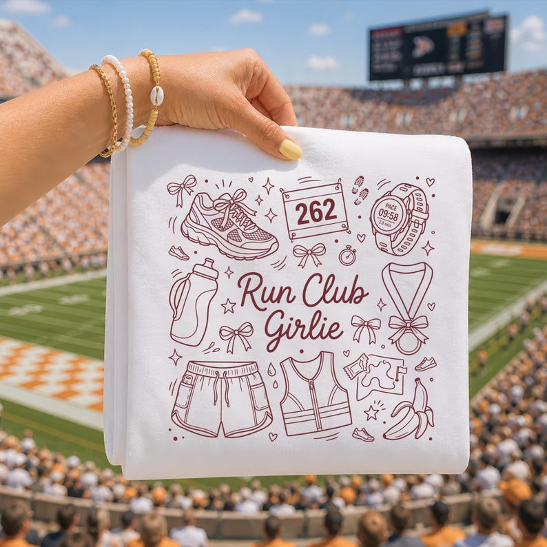 Op de afbeelding: Witte handdoek met een bordeauxrode illustratie van hardloopartikelen, waaronder een schoen, een waterfles, een korte broek en een medaille. De tekst "Run Club Girlie" is ook aanwezig. De handdoek wordt voor een stadion gehouden.