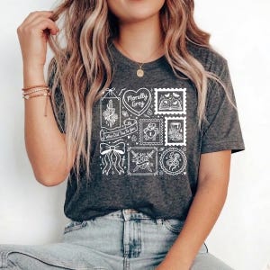 May include: Dark gray t-shirt with a white graphic design featuring various illustrations within stamp-like frames. The design includes a heart, a bow, a sword, and the text "Morally Grey" and "Who Did This To You".