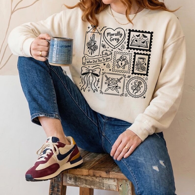May include: Cream-colored sweatshirt with a black graphic design featuring stamps, a heart, a bow, and a sword. The text "Morally Grey" and "Who Did This To You" are also included. The person is wearing blue jeans and sneakers.