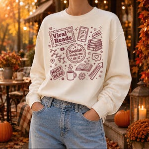 May include: Cream-colored sweatshirt with a maroon graphic design featuring the words "Viral Reads," "Booktok made me do it," and "Must Read." The design includes book-related icons, a coffee cup, and a trending arrow. The person is wearing blue jeans.