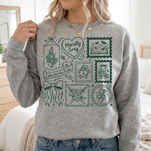 May include: A gray sweatshirt with a green graphic design. The design includes various illustrations such as a heart, a bow, a dagger, and a snake, along with the text "Morally Grey" and "Who Did This To You."