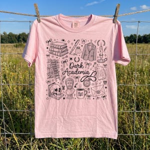 以下が含まれることがあります： 黒いグラフィックデザインの明るいピンク色のTシャツ。デザインには、本、頭蓋骨、胸像、そして「Dark Academia」という言葉が含まれています。その他の要素には、ペン、ジャケット、キャンドル、学術的なイラストが含まれています。